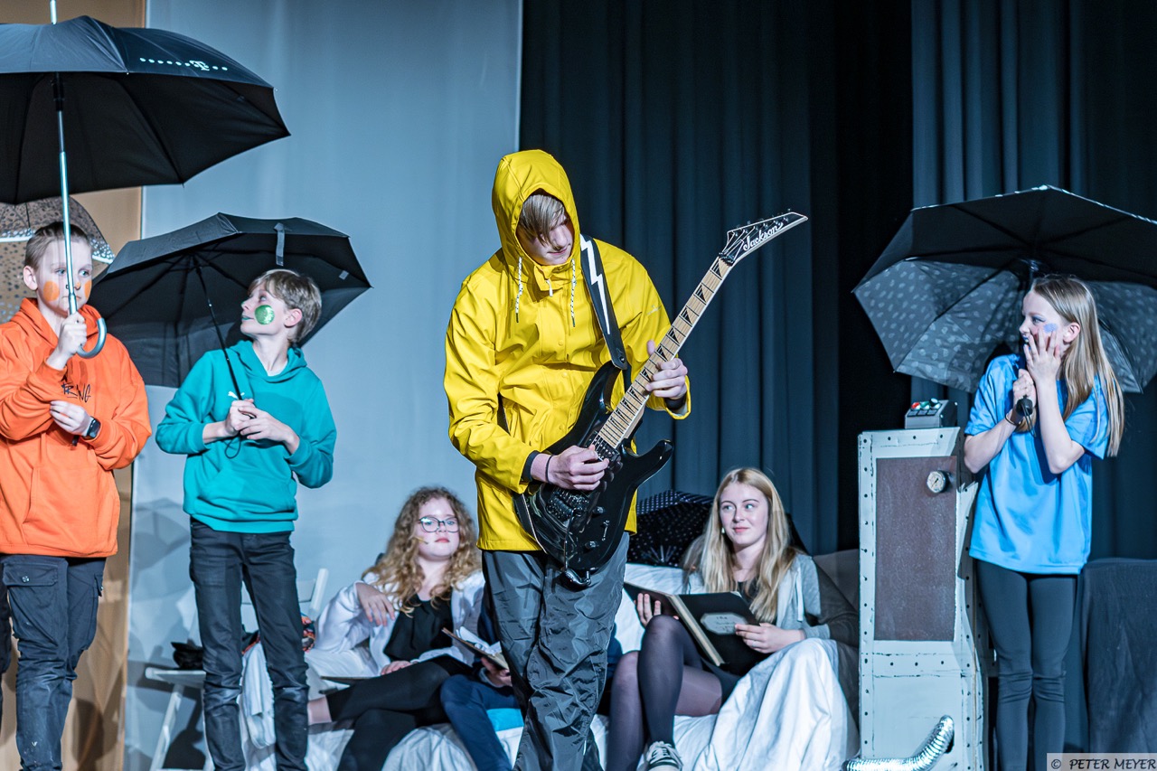 Auf der Bühne steht ein Jugendlicher in einem leuchtend gelben Outfit, der eine E-Gitarre spielt. Um ihn herum befinden sich andere Darsteller mit Regenschirmen und unterschiedlichen Requisiten, die eine vielfältige, dynamische Szene darstellen.